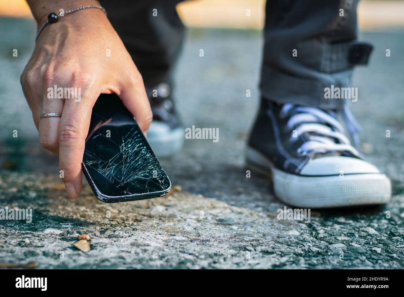 Donna che raccoglie lo smartphone rotto da terra. Telefono cellulare danneggiato con schermo a sfioramento incrinato. Foto Stock