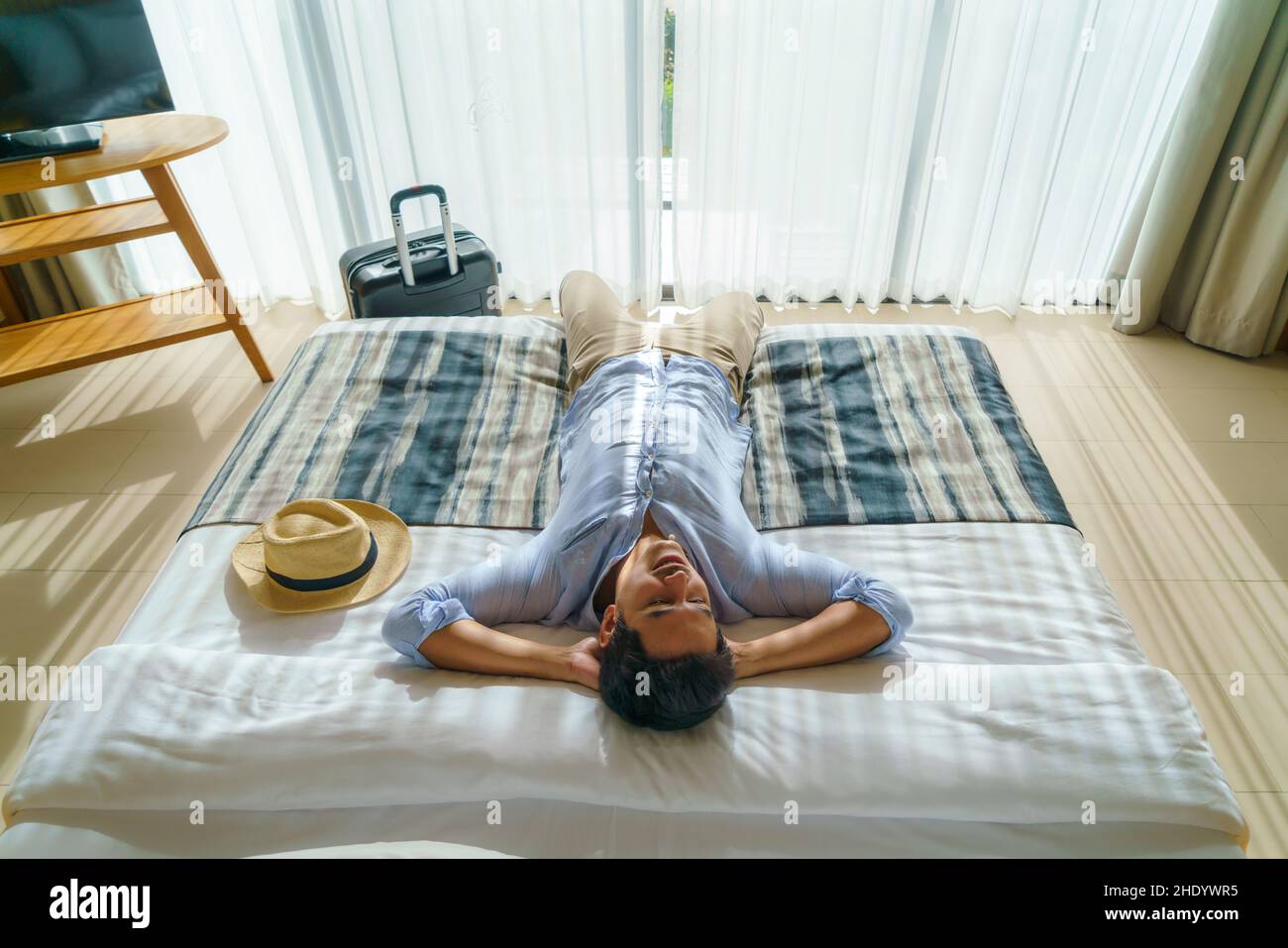 L'uomo asiatico con una valigia si trova su un letto nella sua camera in resort dopo il suo arrivo durante la sua vacanza estiva viaggio. Foto Stock