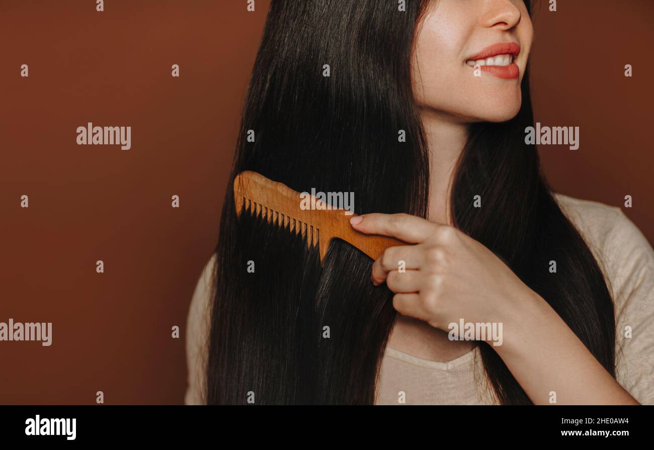 Giovane donna bruna felice pettinando i suoi lunghi capelli sani marrone e sorridendo alla macchina fotografica mentre si posa isolato su sfondo scuro con spazio copia per il testo Foto Stock