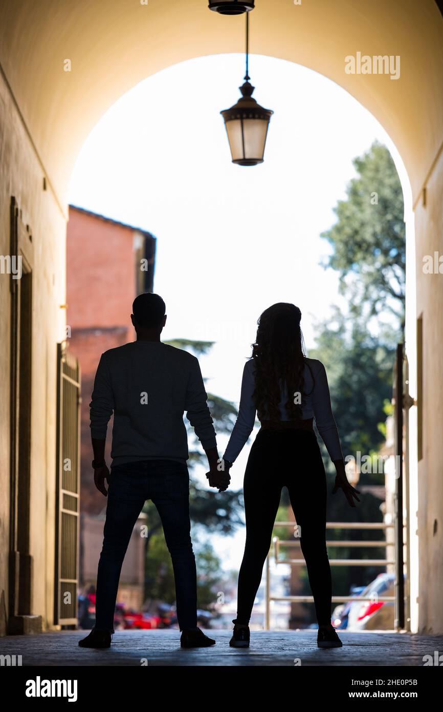 Silhouette umane in arca antico con lampada da strada appesa sopra. L'uomo e la donna si tengono a mano, girati indietro, alberi e case dentro Foto Stock