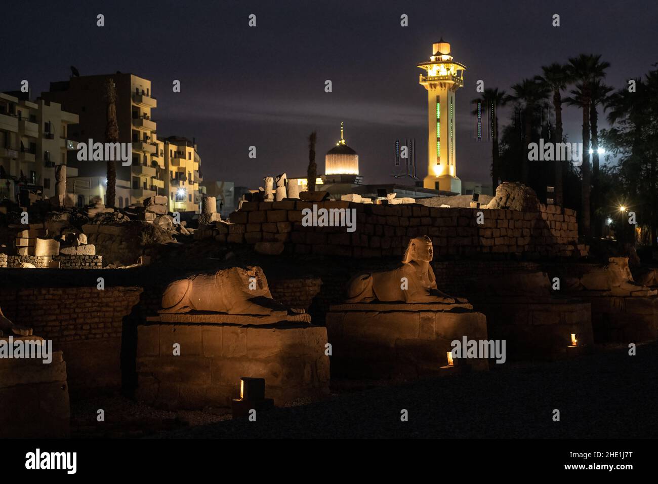Statue Sfinge che costeggiano la strada dei arieti a Luxor, egitto, con la città e il minareto illuminato dietro lo sfondo. Foto Stock