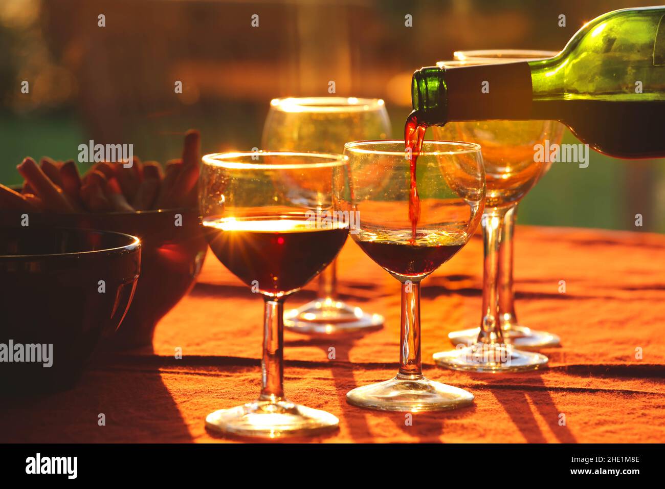 Versare il vino rosso dalla bottiglia in bicchieri sul tavolo con spuntini sullo sfondo durante il tramonto Foto Stock