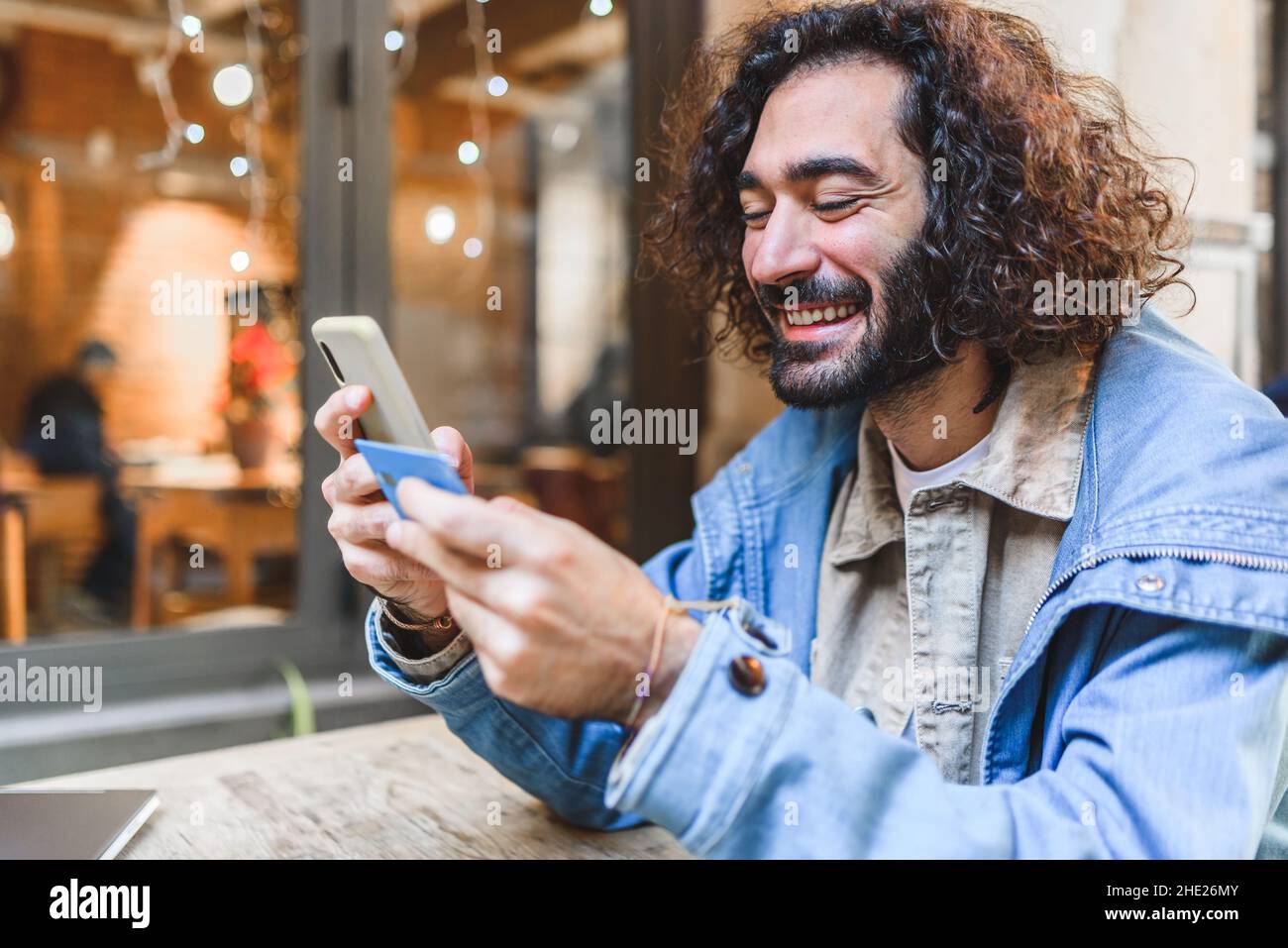 Maschio ispanico gleful in abiti casual con capelli ricci e barba sorridente. Sta inserendo le credenziali della carta di credito nel cellulare mentre si siede al tavolo della caffetteria e shopping in linea sulla strada Foto Stock