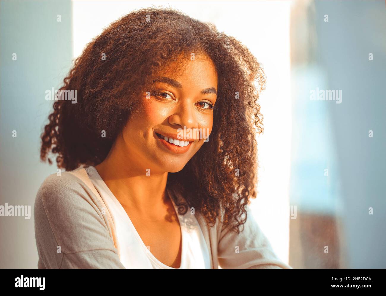 Felice African American ragazza etnia rilassato in abiti da casa seduti su davanzale sullo sfondo di tende che ondolano dal vento chiaro, felice afro Foto Stock