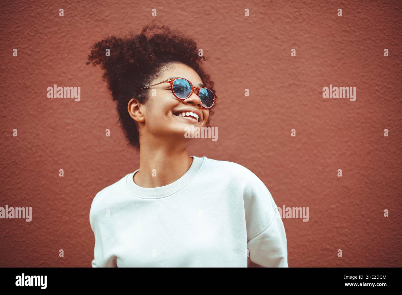 Etnia africana giovane donna in occhiali da sole eleganti, con capelli ricci legati in alto ponytail, guardando via, mentre sorridendo ampiamente mostrando dritto per Foto Stock