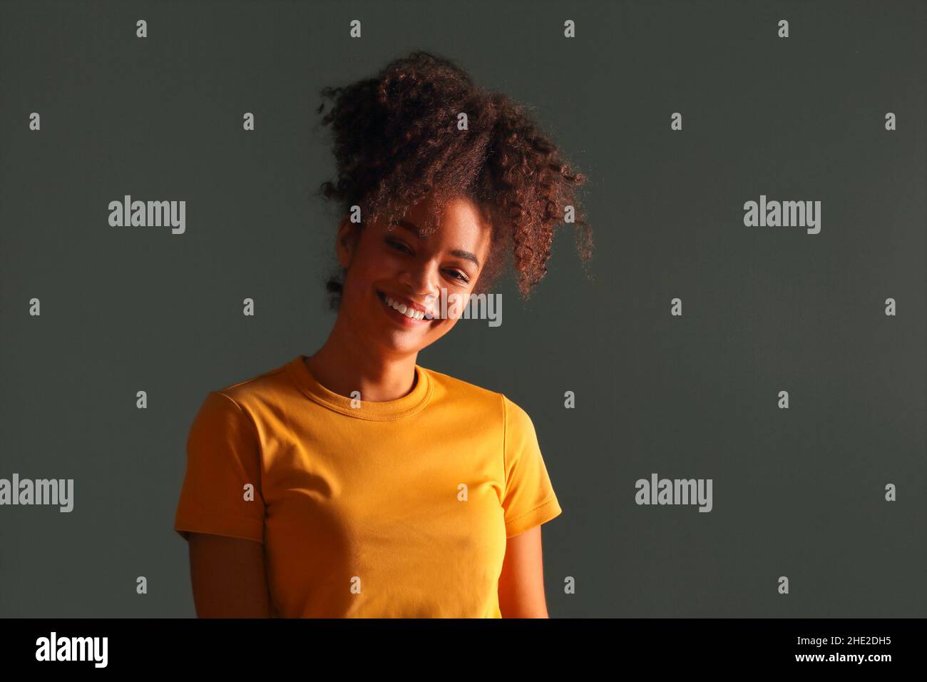 Bella ragazza di etnia afroamericana felice scuro con pelle in maglia gialla che alza la mano nel saluto mentre guarda in macchina fotografica con piacevole sorriso, p Foto Stock