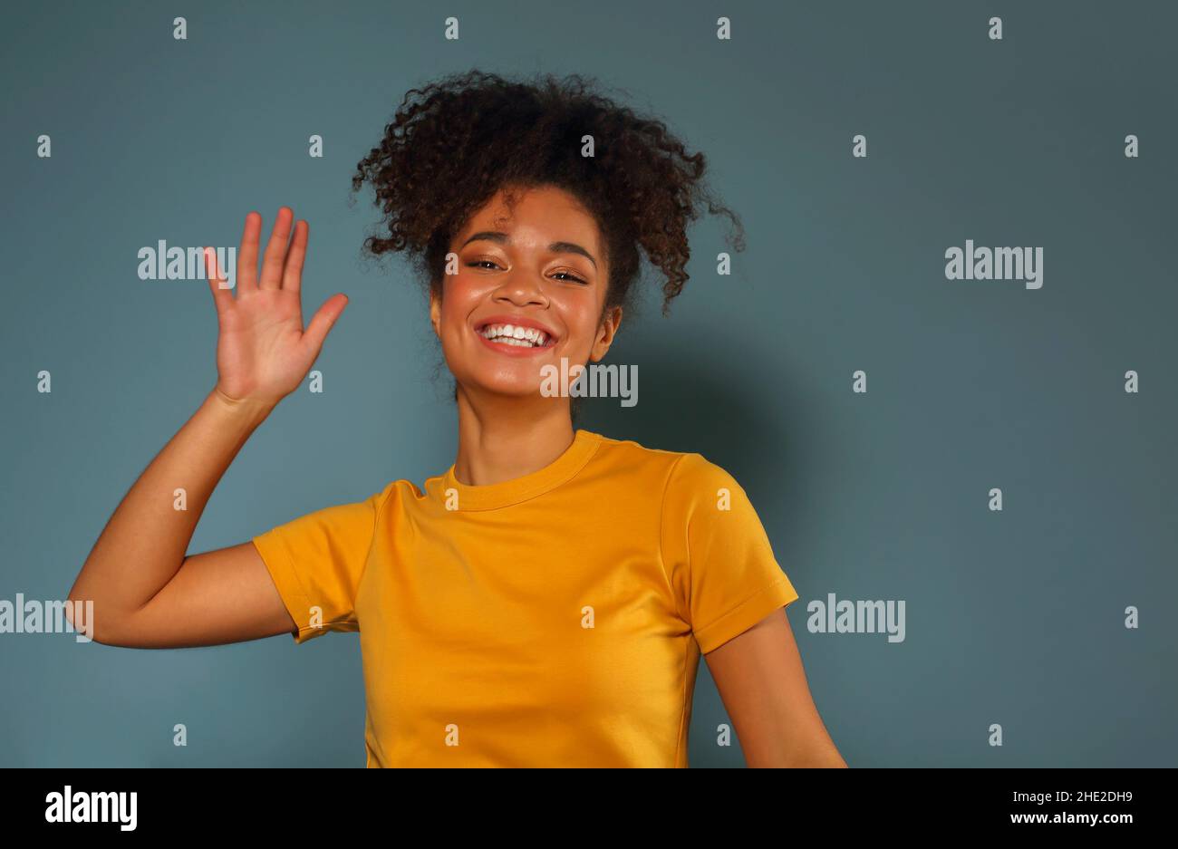 Bella ragazza di etnia afroamericana felice scuro con pelle in maglia gialla che alza la mano nel saluto mentre guarda in macchina fotografica con piacevole sorriso, p Foto Stock