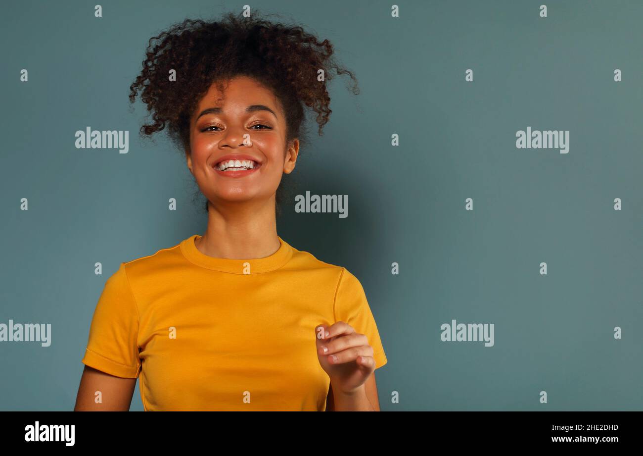 Bella ragazza di etnia afroamericana felice scuro con pelle in maglia gialla che alza la mano nel saluto mentre guarda in macchina fotografica con piacevole sorriso, p Foto Stock