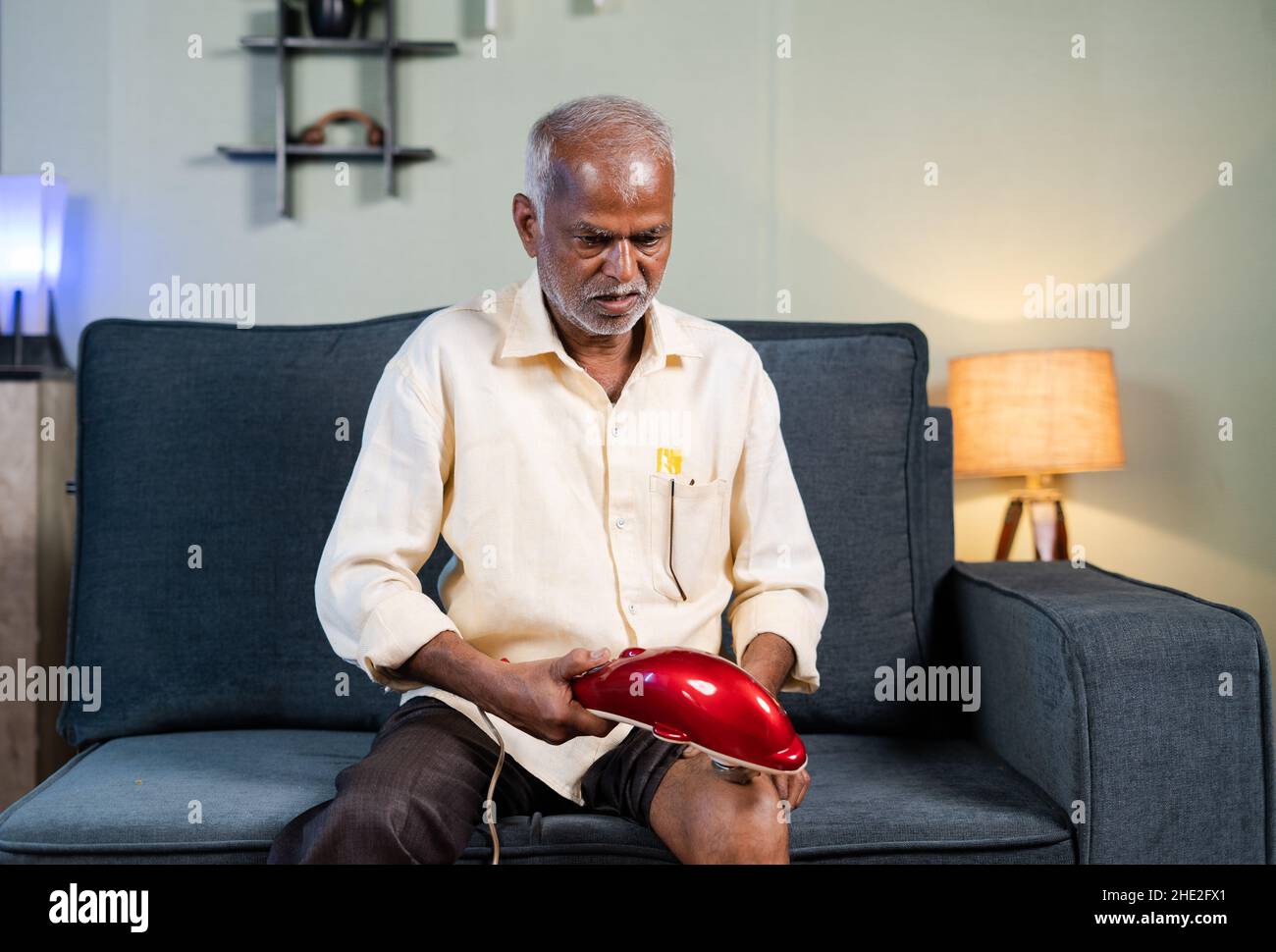 Uomo anziano che usa la macchina di massaggio di vibrazione per rilievo di dolore dell'articolazione del ginocchio nel paese sul divano - concetto di macchina elettronica di rilievo di dolore e terapia o. Foto Stock