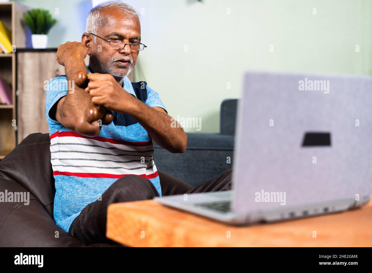 uomo anziano indiano massaggiante mano utilizzando il massaggiatore a rullo di legno guardando i video online dei farmaci sul portatile da casa - concetto di fisioterapia Foto Stock