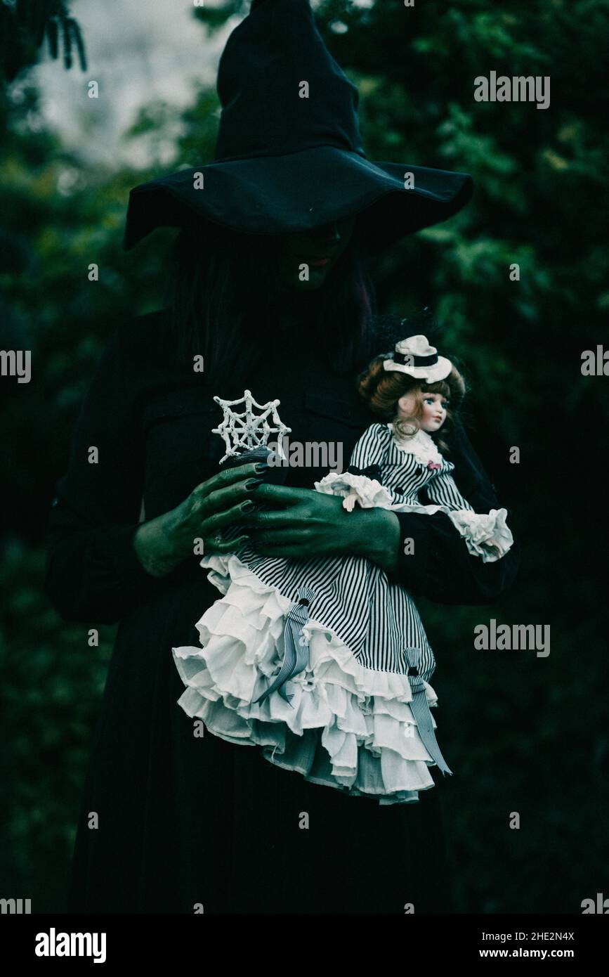 Scatto verticale di una bella giovane strega con un costume di Halloween e bambola in giardino Foto Stock