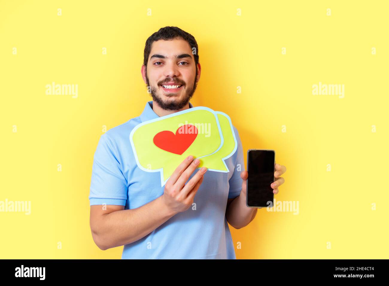 Il giovane uomo tiene in mano un fumetto con un cuore e mostra lo schermo vuoto dello smartphone. Concetto di social media. Foto Stock