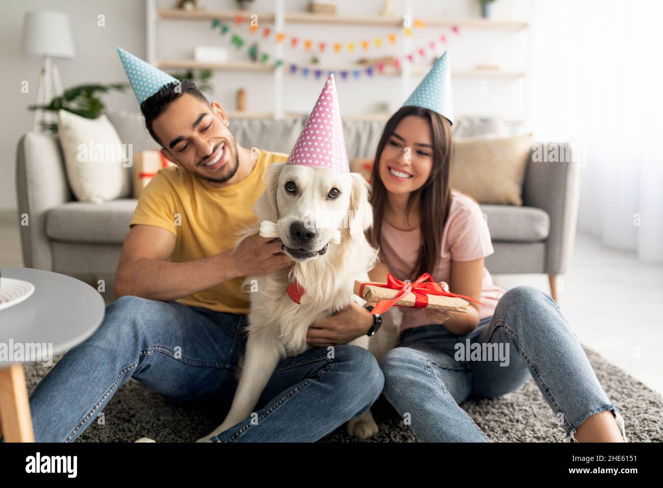 Coppia multirazziale millennial in cappelli da festa che tengono la scatola del regalo, celebrando il compleanno del cane dell'animale domestico nel paese Foto Stock