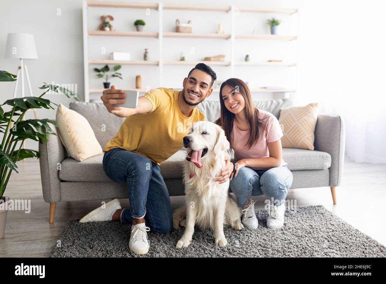 Positivo giovane coppia diversificata prendere selfie con il loro cane mentre si siede sul pavimento a casa, a tutta lunghezza Foto Stock