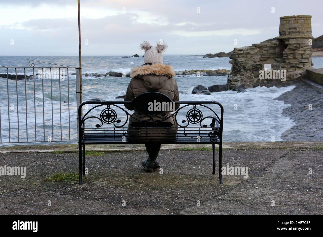 Dunure, Ayrshire, Scozia, Regno Unito. Una donna di giorno di inverni si siede su una panchina con la schiena alla macchina fotografica che indossa un cappello di bobble guardando il mare tempestoso Foto Stock