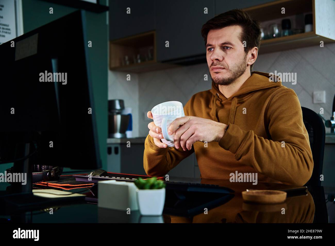 L'uomo lavora in ufficio a casa con una tazza di caffè in mano, usa il computer. Freelance che lavora in remoto in ritardo. Stress emotivo e burnout Foto Stock