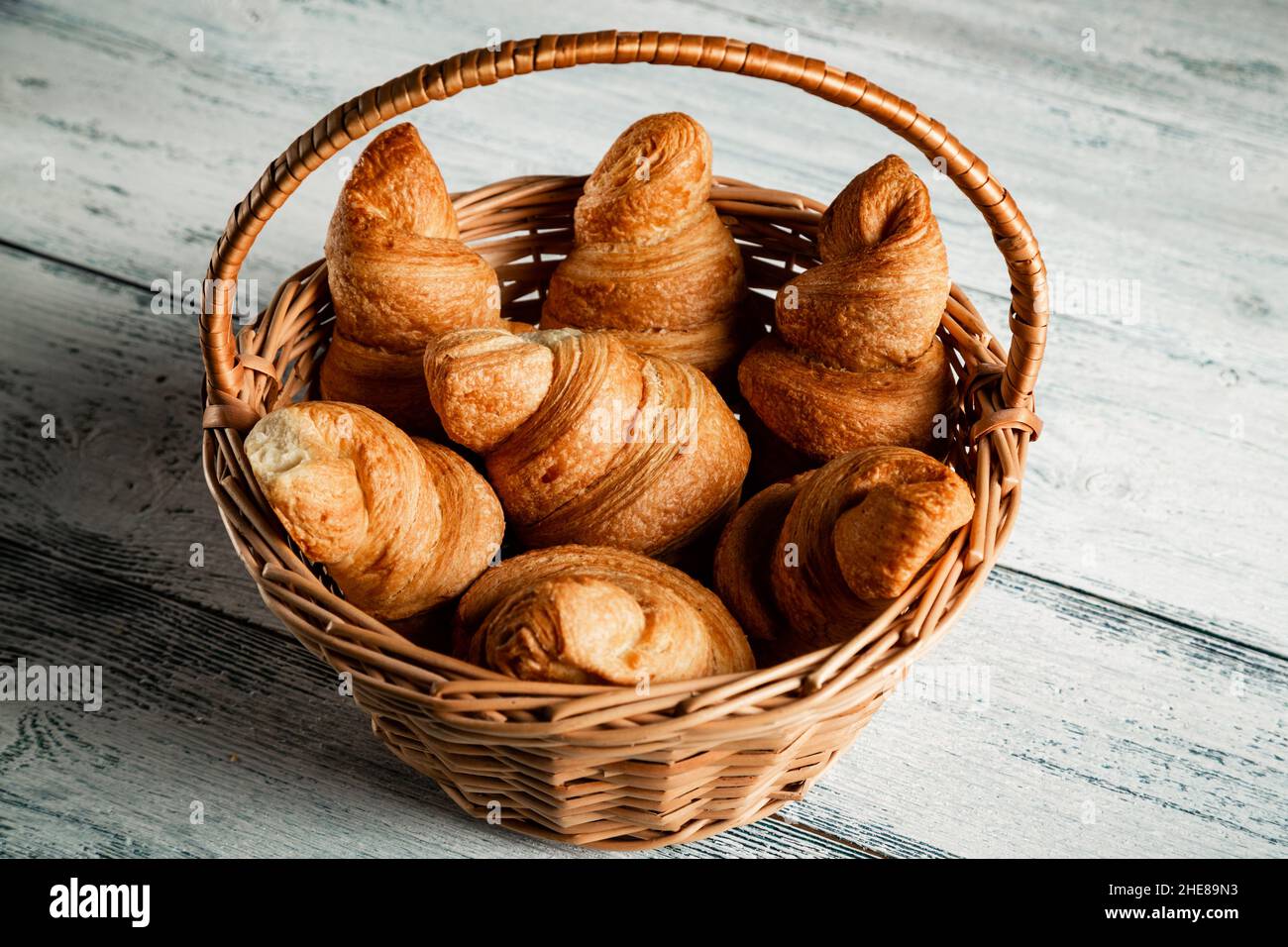 Croissant in un cestino di vimini su sfondo di legno. Vecchie tavole bianche e blu. Foto Stock