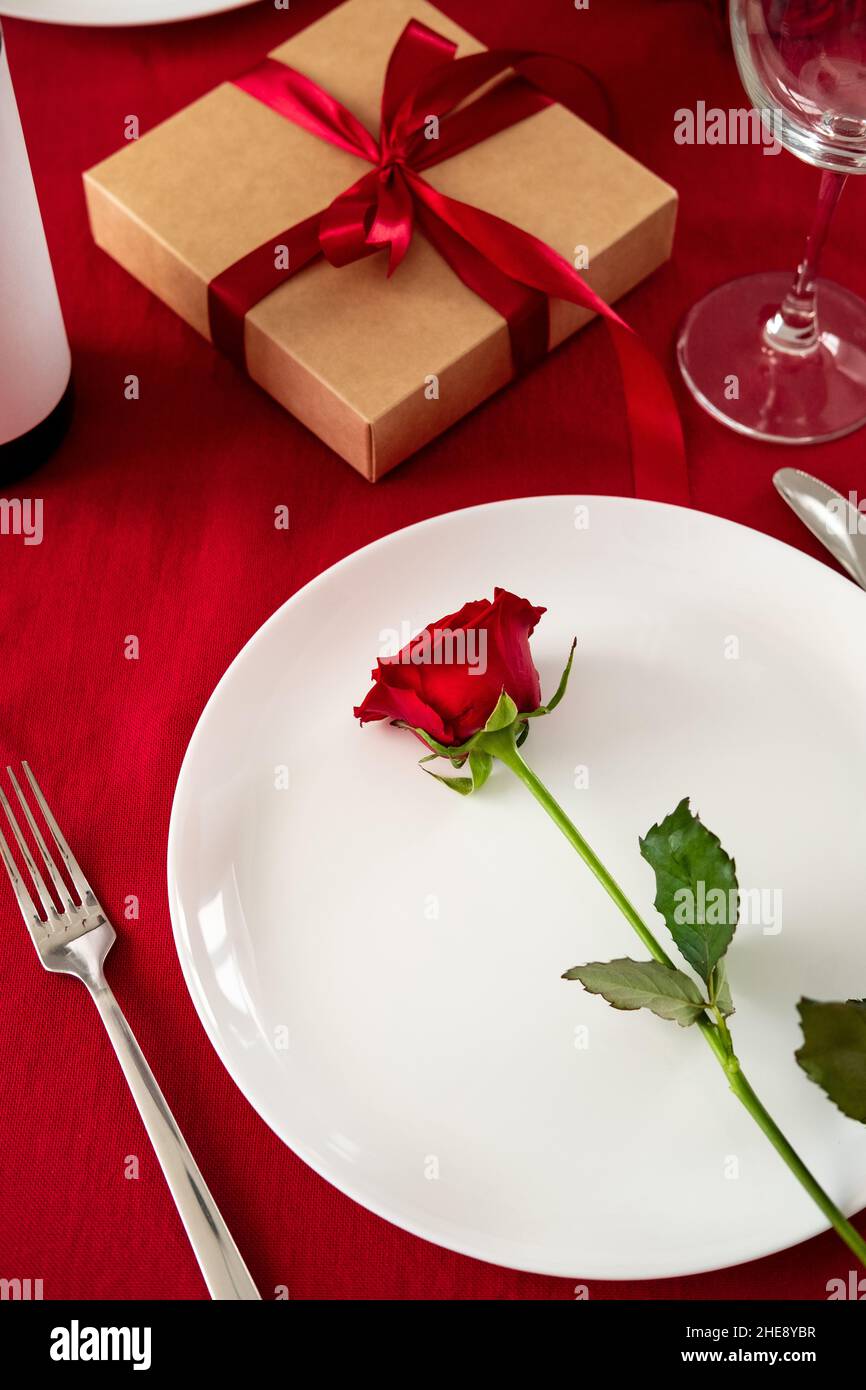 Cena di San Valentino. Rosa su piatto bianco, scatola regalo, posate, vino, bicchieri su sfondo rosso Foto Stock