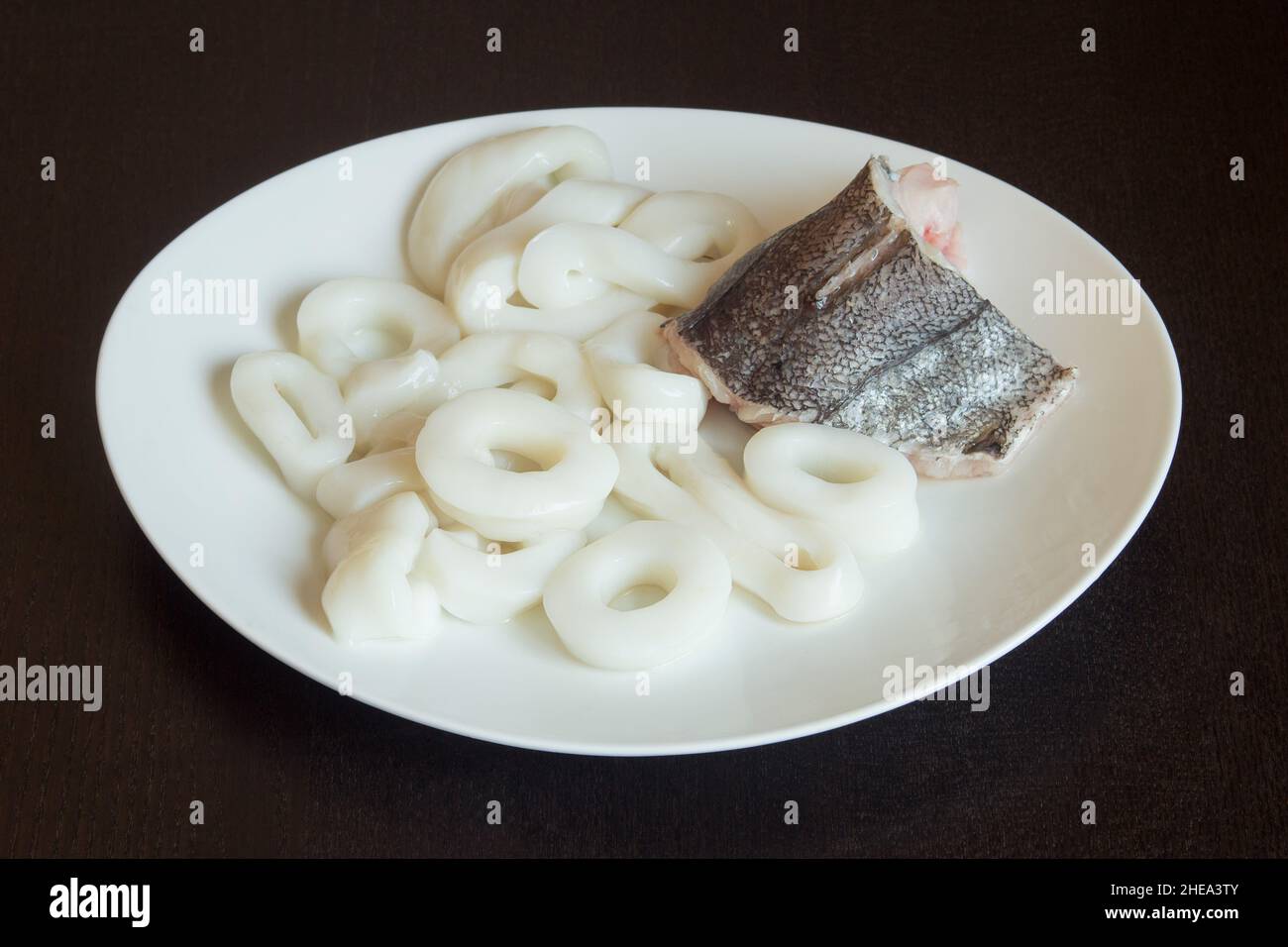 Un piatto bianco su un tavolo di legno con un pezzo di nasello fresco con anelli di calamari crudi. Cucina sana e pesce. Foto Stock