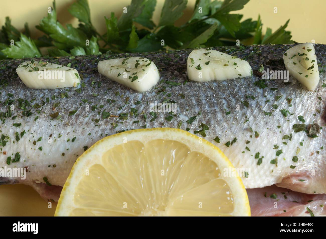 Pezzi di aglio conditi con spezie in fila sulla pelle di branzino crudo accompagnato da ciuffi di prezzemolo fresco e una fetta di limone. Preparazione del pesce. Foto Stock