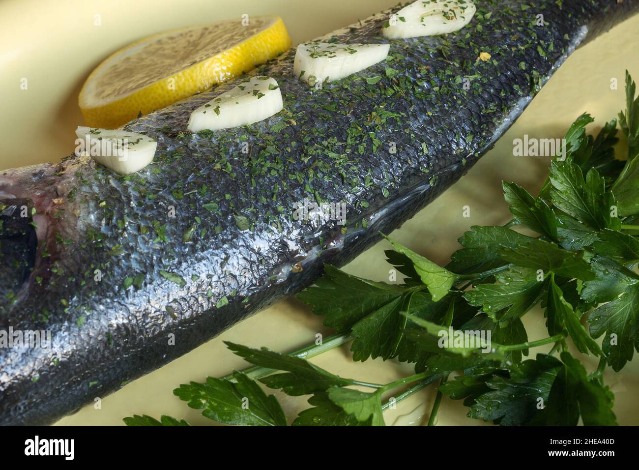 Rametti di prezzemolo fresco e specie secche e schiacciate su spigola cruda in un piatto di ceramica dipinta di giallo e un cuneo di limone. Cibo e pesce sani. Foto Stock