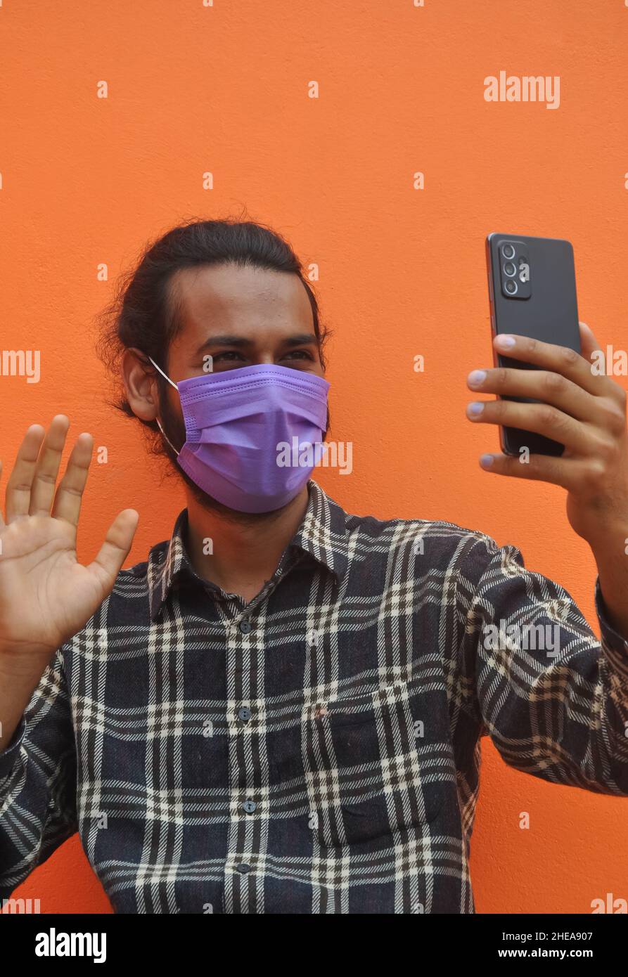 Primo piano di un giovane ragazzo che indossa la maschera facciale, che dice ciao con ondeggiare la mano a qualcuno mentre videochiama, posando contro il muro arancione. Foto Stock