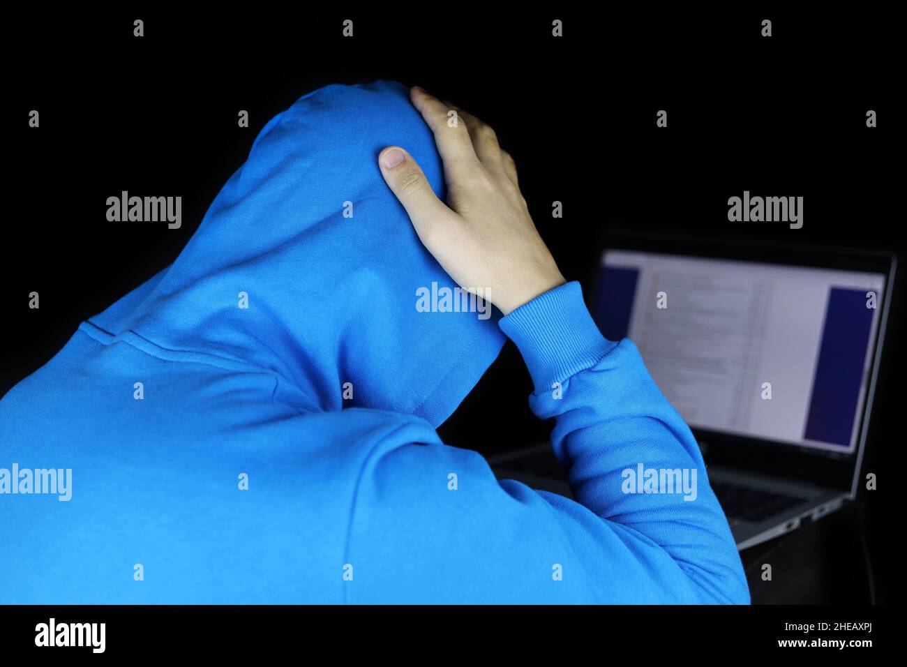 Uomo con cappuccio blu seduto con un computer portatile che gli aggrappa la testa. Concetto di cybercriminalità, hacking, compito difficile o errore fatale Foto Stock