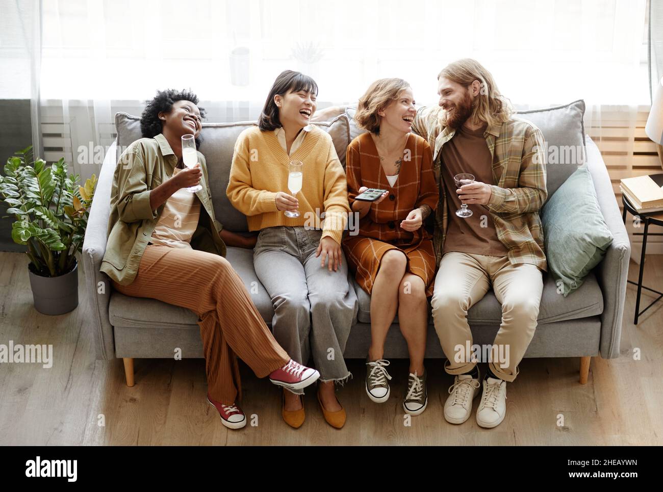 Gruppo di amici felici seduti sul divano bere champagne e ridere durante la conversazione in camera Foto Stock