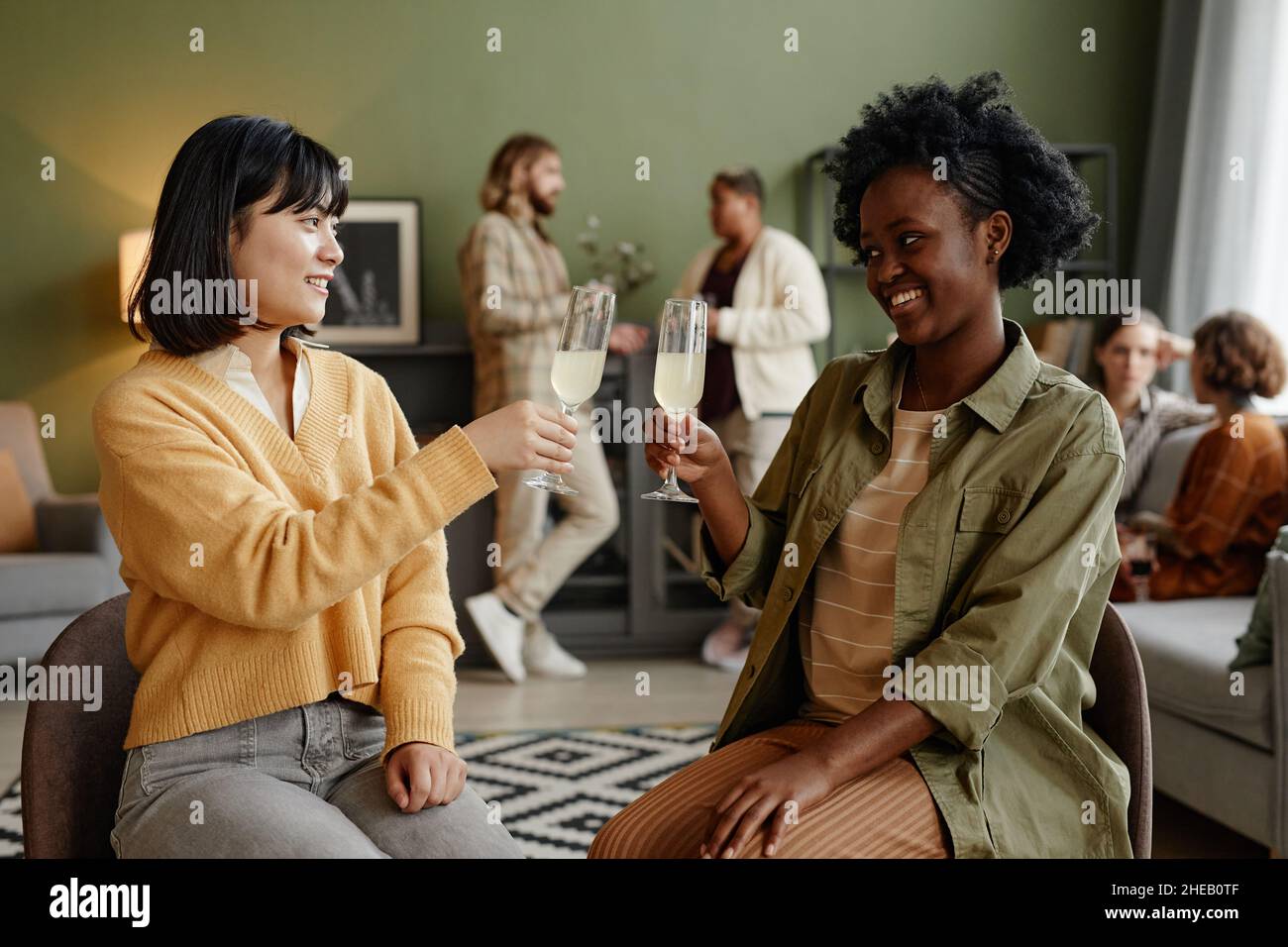 Le giovani donne tostano con flute di champagne mentre si siedono a festa in soggiorno con altre persone sullo sfondo Foto Stock