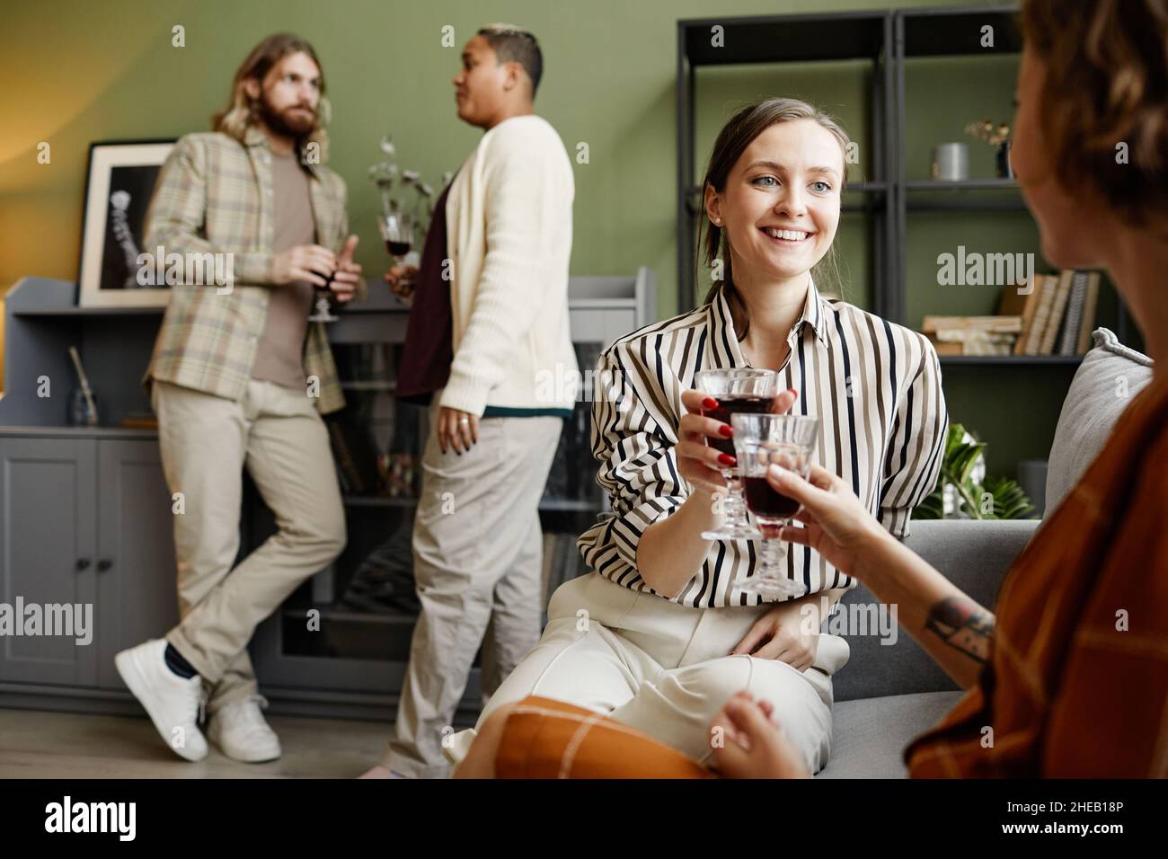 Giovani donne felici che tostano con bicchieri di vino rosso mentre si siedono sul divano durante la festa di casa con altre persone sullo sfondo Foto Stock