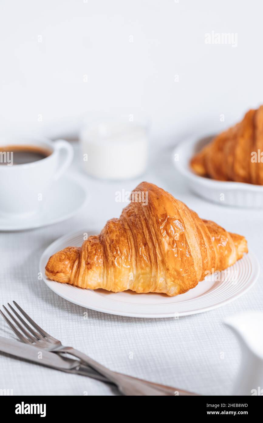 Croissant in un ambiente bianco luminoso, con una tazza di caffè e latte e altri croissant in uno sfondo sfocato. Ambiente colazione semplice ed elegante Foto Stock
