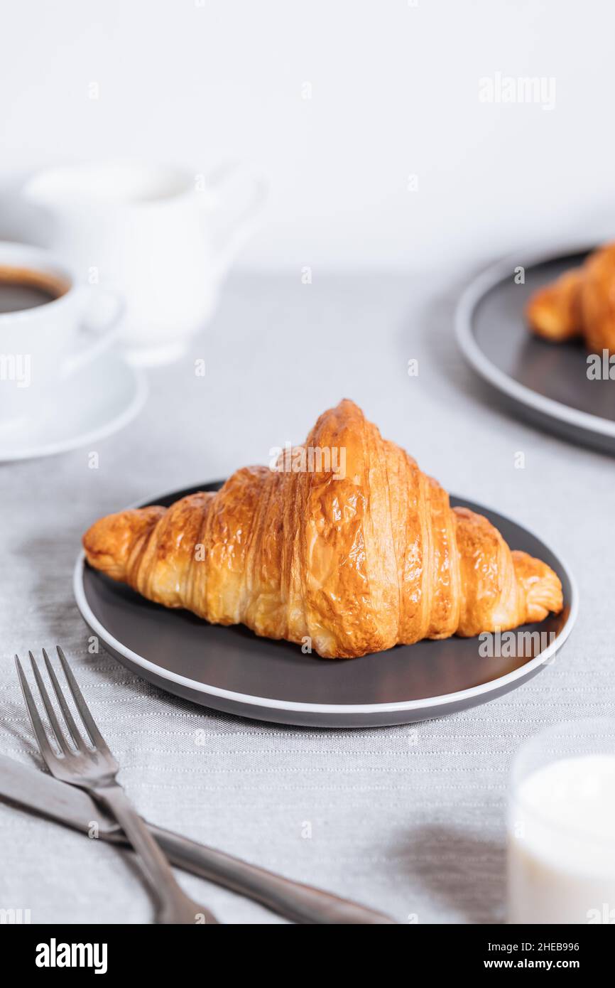 Croissant in un ambiente bianco luminoso, con una tazza di caffè e latte e altri croissant in uno sfondo sfocato. Ambiente colazione semplice ed elegante Foto Stock