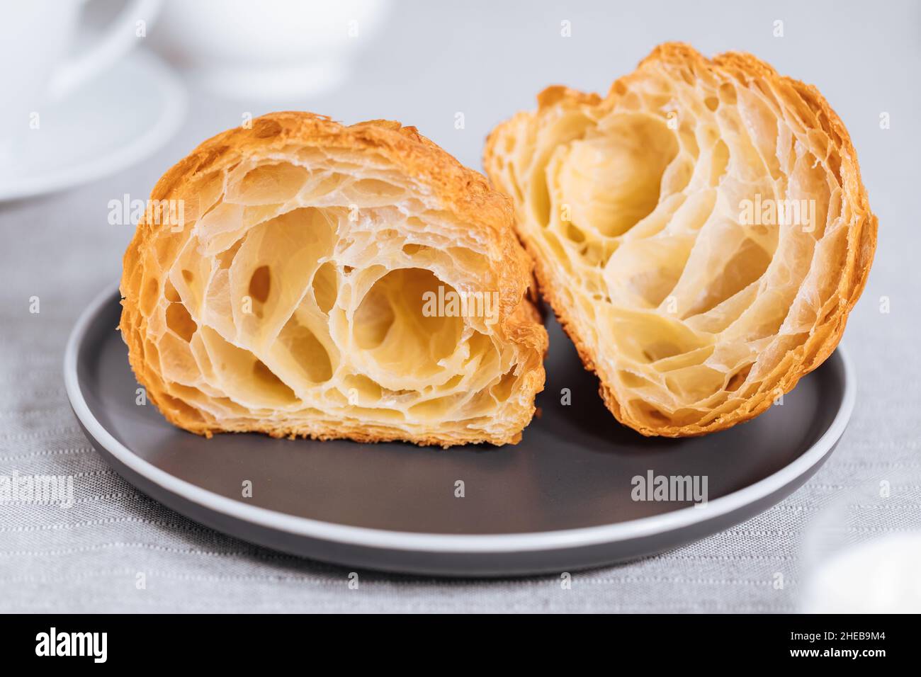 Croissant in un ambiente grigio chiaro e tagliato a due pezzi, con una tazza di caffè e latte e altri croissant in uno sfondo sfocato. Semplice ed elegante b Foto Stock