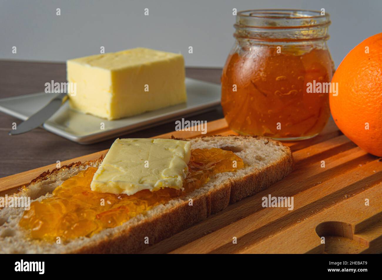 pane rustico con marmellata d'arancia e burro su un asse di legno con un vasetto di marmellata sullo sfondo accanto al burro Foto Stock