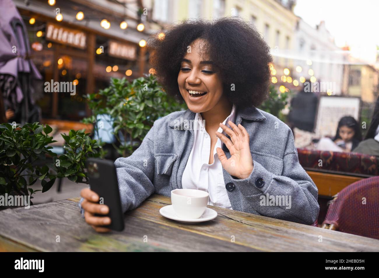 Ritratto Street style di attraente giovane felice afroamericana donna con capelli afro hanno video chat con smartphone all'aperto in caffè marciapiede. Eccitata sorridente femmina blogger trasmette live streaming Foto Stock