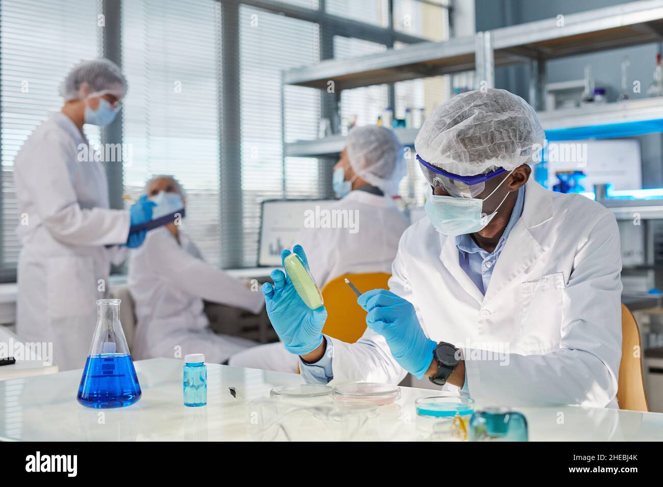 Scienziato africano in abbigliamento protettivo che prende il campione e lo esamina durante l'esperimento in laboratorio con i suoi colleghi in background Foto Stock