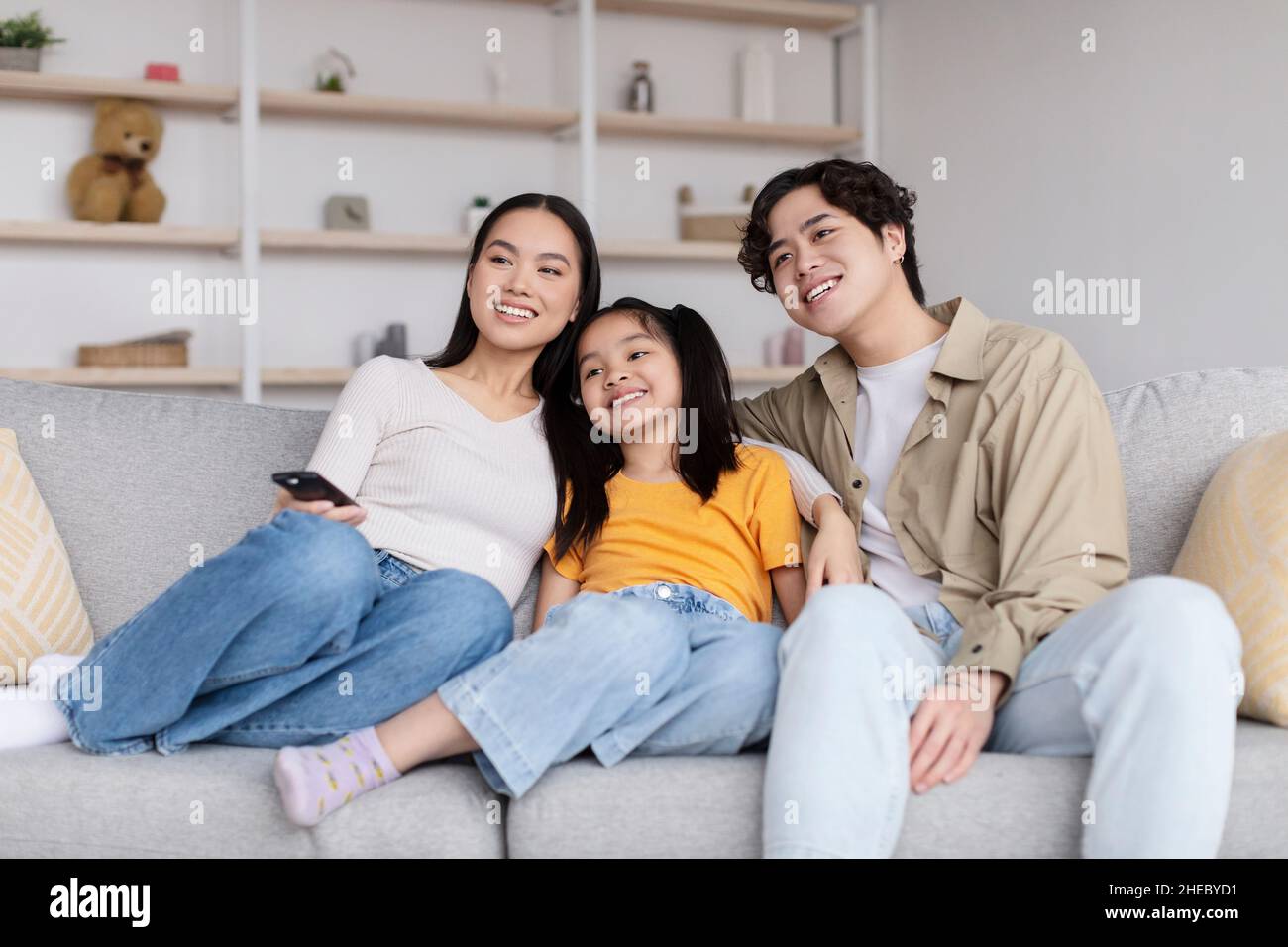 Sorridente madre coreana, padre e ragazza adolescente abbracciano e guardano la tv, siediti sul divano in soggiorno Foto Stock