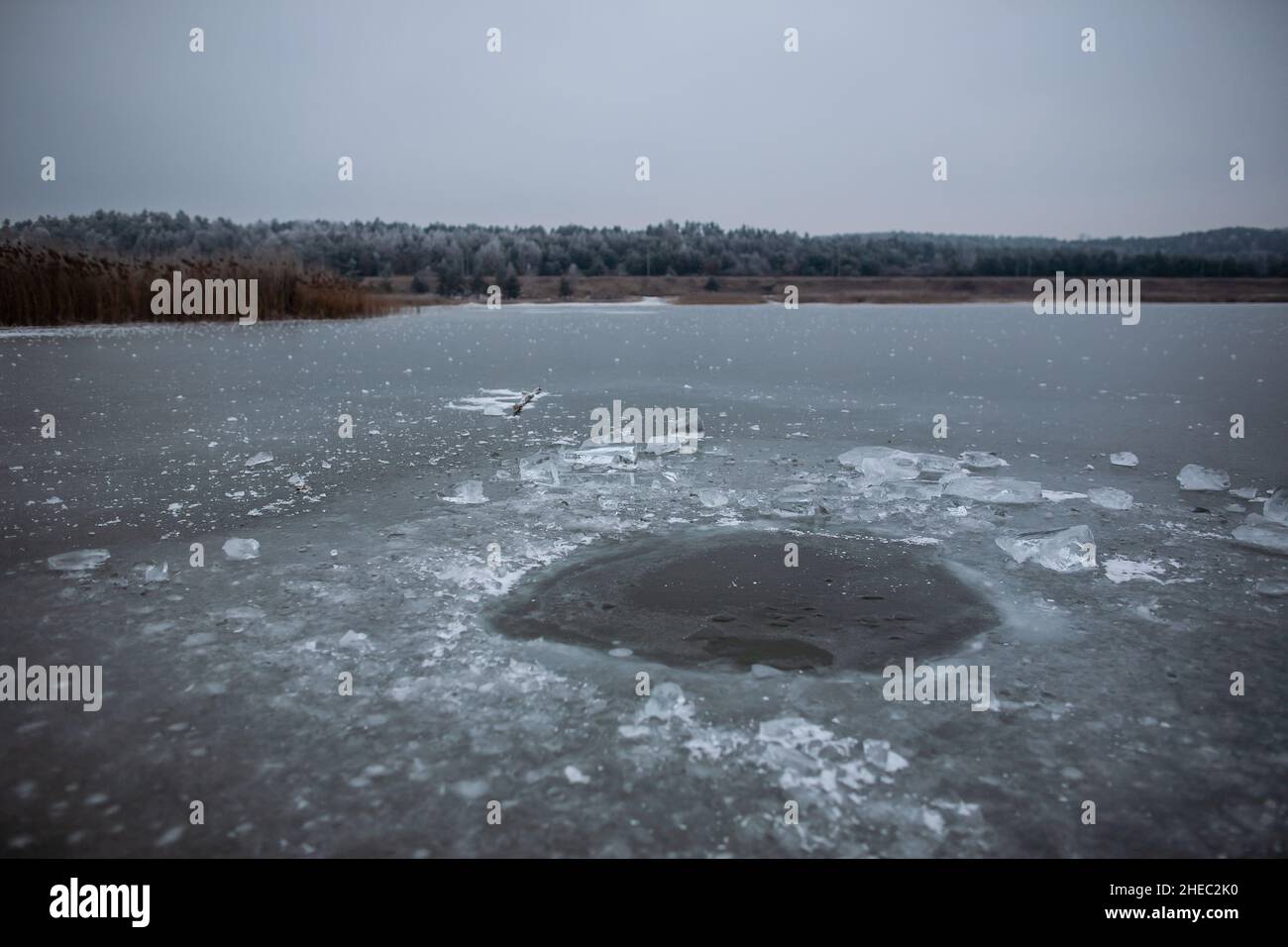 Buca in ghiaccio sulla superficie dell'acqua con blocchi di ghiaccio sparsi intorno in giorno nuvoloso, canne e foresta in background, ghiaccio sul lago congelato Foto Stock