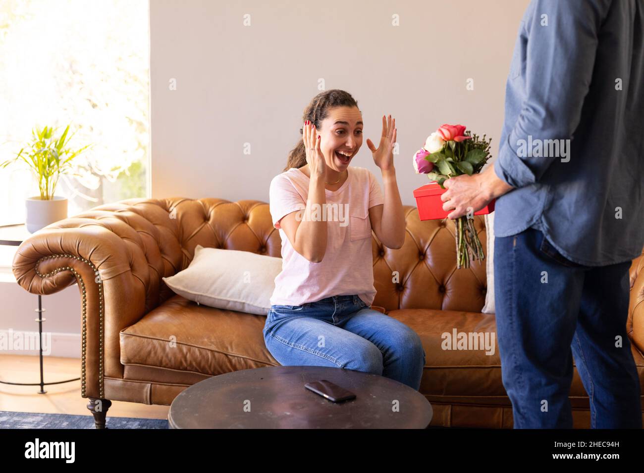 Metà sezione di un uomo che sorprende sua moglie con un bouquet di fiori e scatola regalo a casa Foto Stock