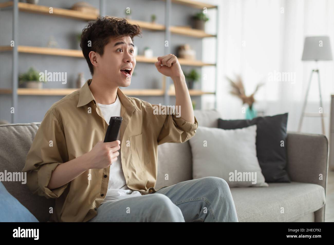 Ragazzo asiatico eccitato che trascorre il fine settimana guardando la tv scuotendo pugno clenched Foto Stock