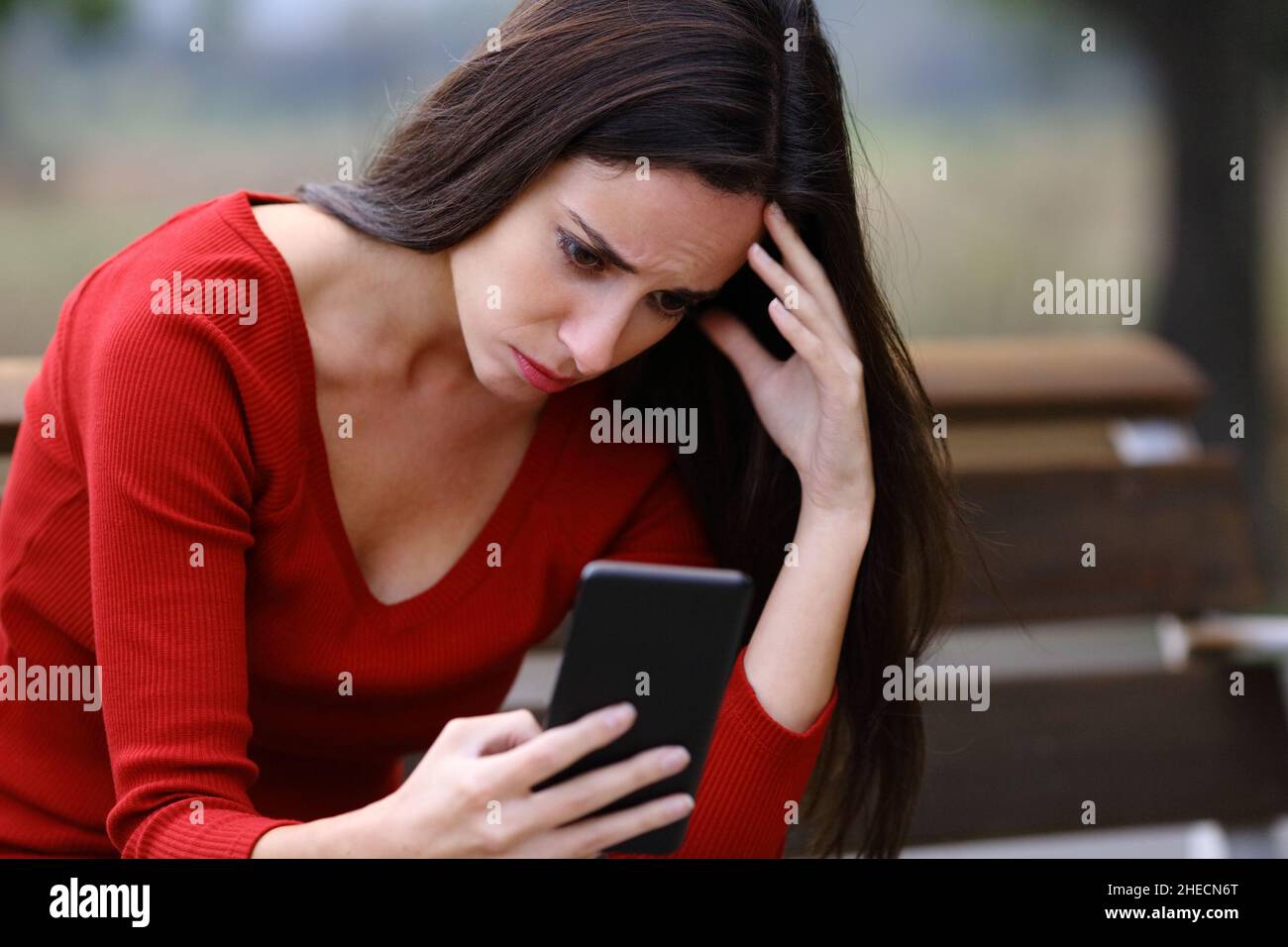 Donna preoccupata seduta su una panca che controlla il contenuto dello smartphone in un parco Foto Stock