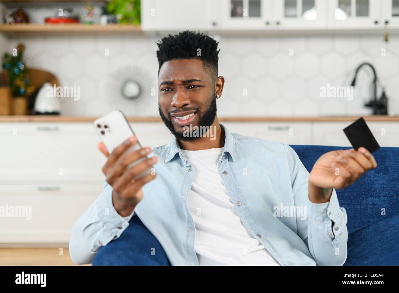 Afro-americani preoccupati per il denaro. Confuso uomo nero in possesso di carta di credito e smartphone con volto deluso, shrugging, fallimento concetto Foto Stock
