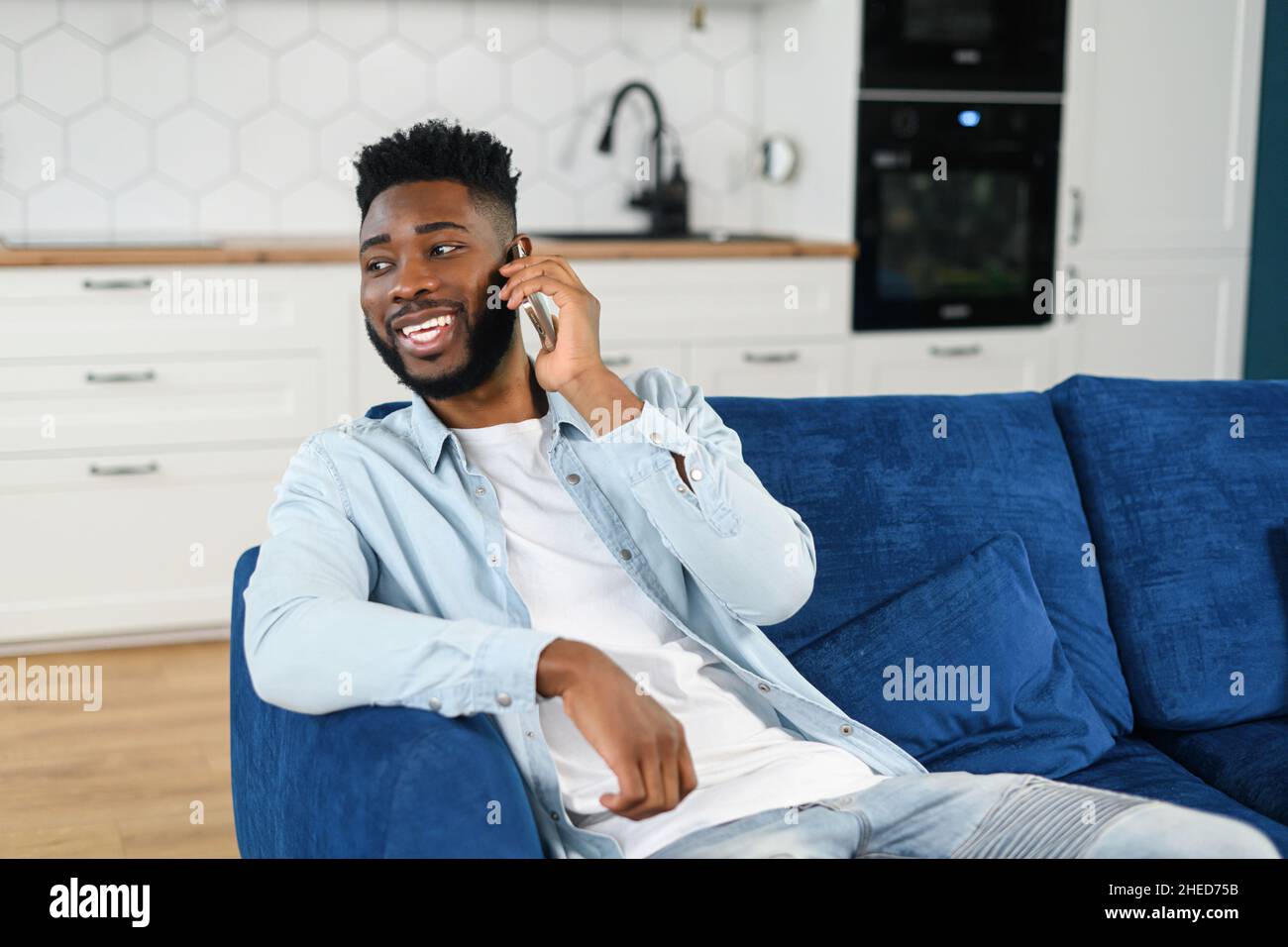 Felice bel ragazzo afroamericano ha divertente conversazione telefonica seduta sul divano a casa, ragazzo biraciale overjoyed che parla sullo smartphone, chiacchierando sul telefono cellulare, si sente felice e gioia Foto Stock
