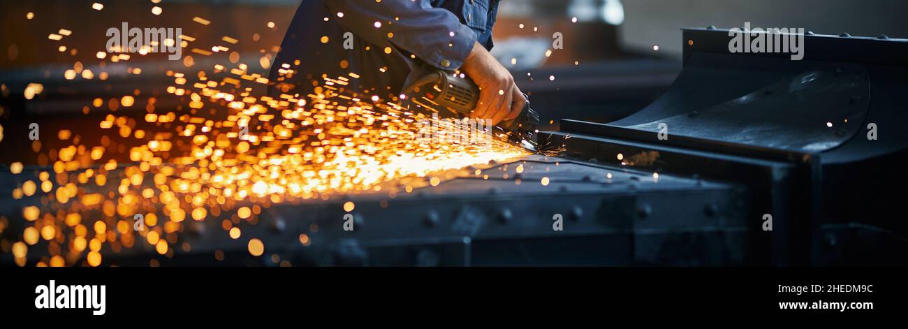 Primo piano di lavoro in uniforme blu scuro che tiene speciali attrezzature per lucidare il metallo in smithy. Concetto di processo che rende moderno recinto vicino a scintille arancioni. Foto Stock