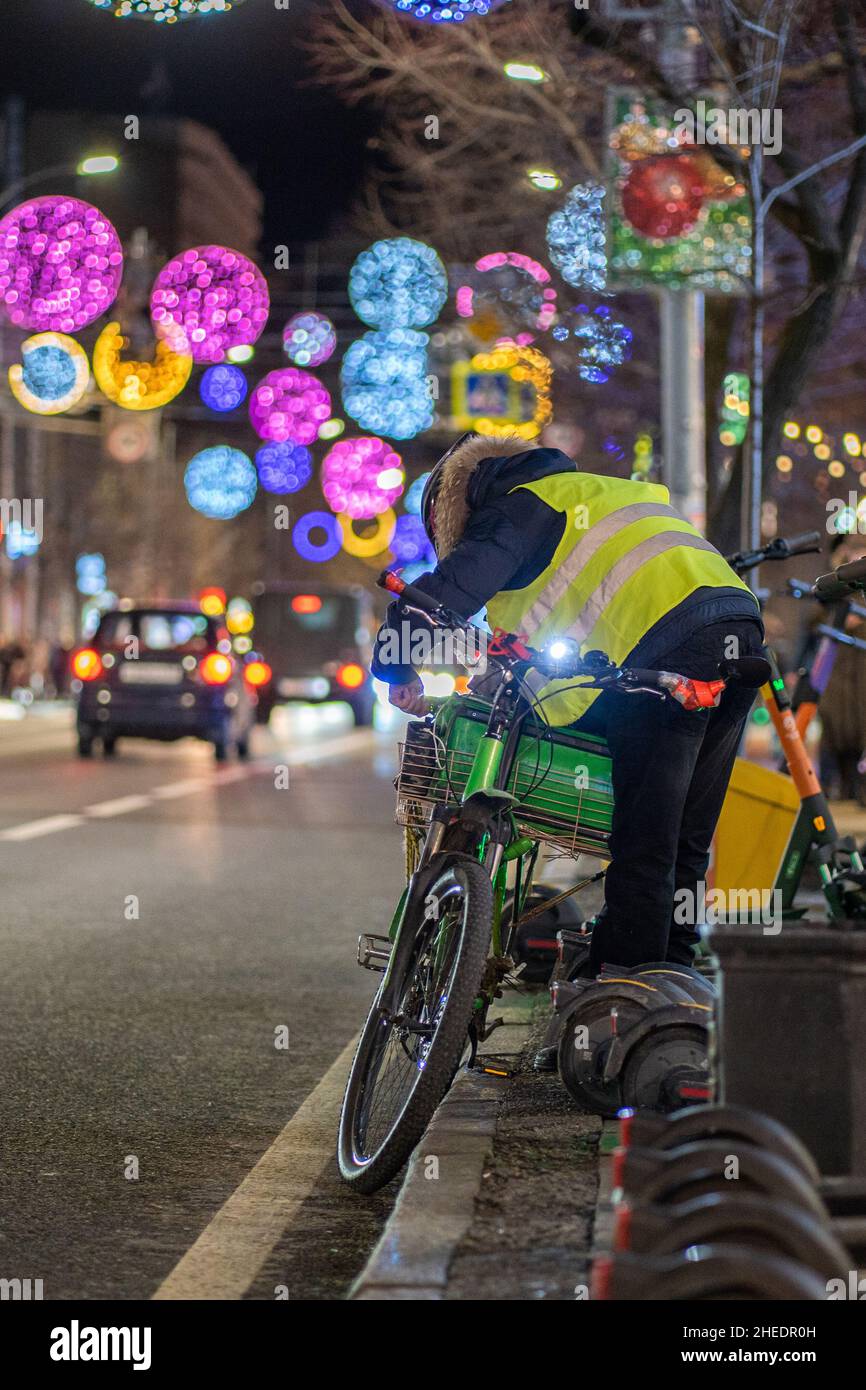 L'addetto alla consegna degli alimenti fissa il sacchetto termico al portabiciclette. L'uomo che indossa un giubbotto di sicurezza con strisce riflettenti si trova sul bordo della strada Foto Stock