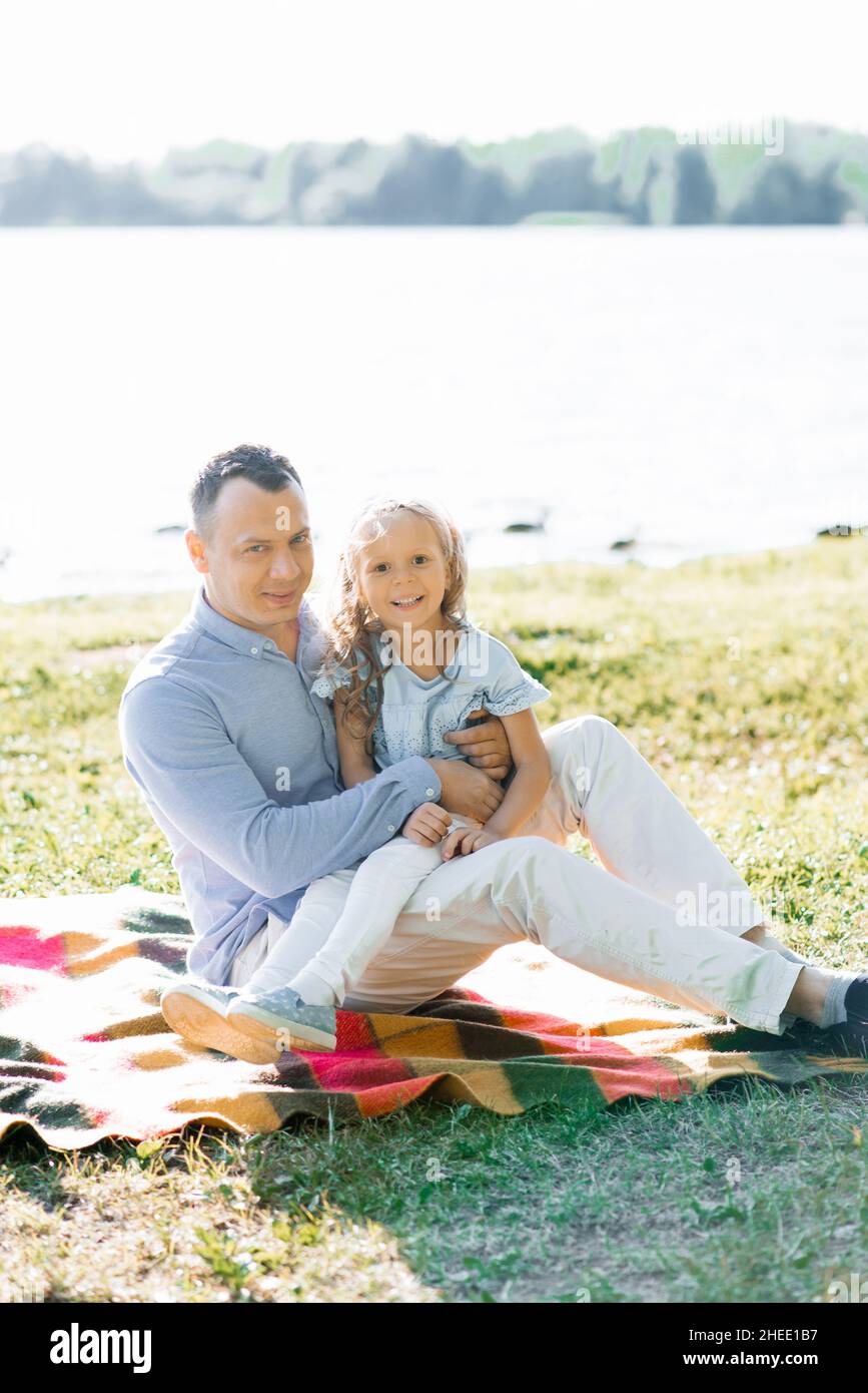 Un padre felice tiene la figlia tra le braccia. Si divertono in natura, seduti su una coperta vicino al lago in estate Foto Stock