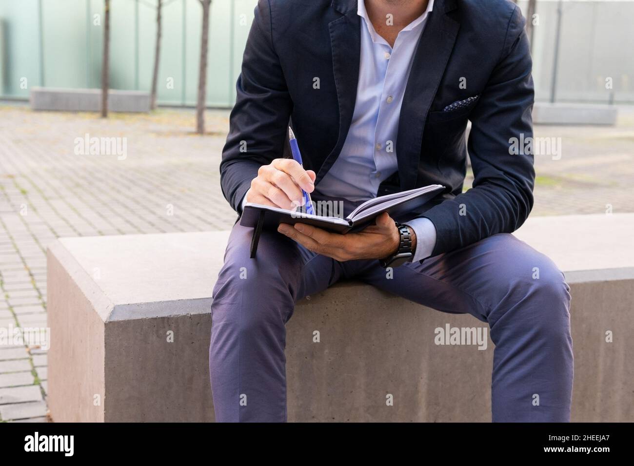 Ritagliare l'imprenditore maschio senza volto in abito elegante prendendo appunti in blocco appunti mentre si siede sul bordo di pietra sulla strada in città Foto Stock