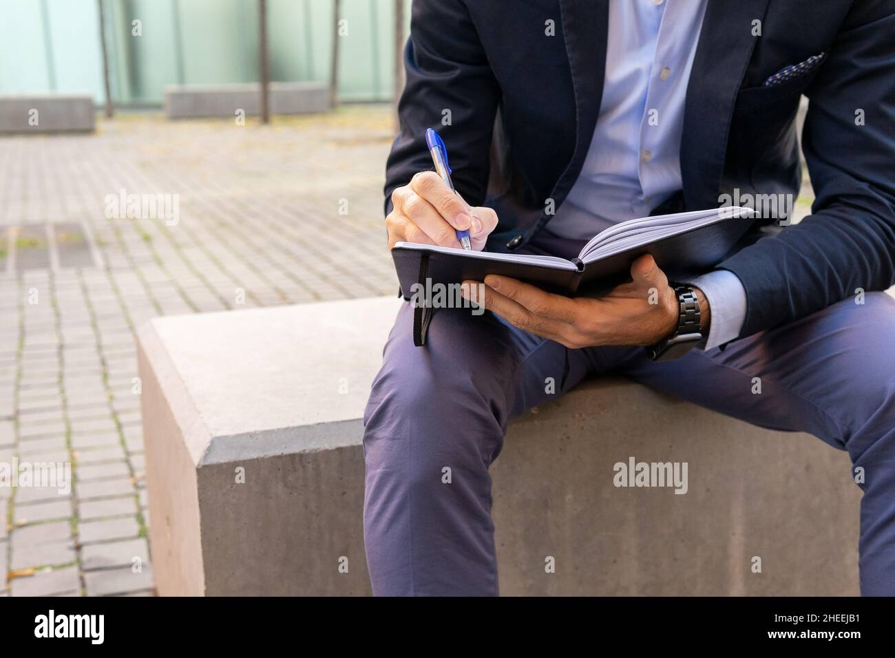 Ritagliare l'imprenditore maschio senza volto in abito elegante prendendo appunti in blocco appunti mentre si siede sul bordo di pietra sulla strada in città Foto Stock