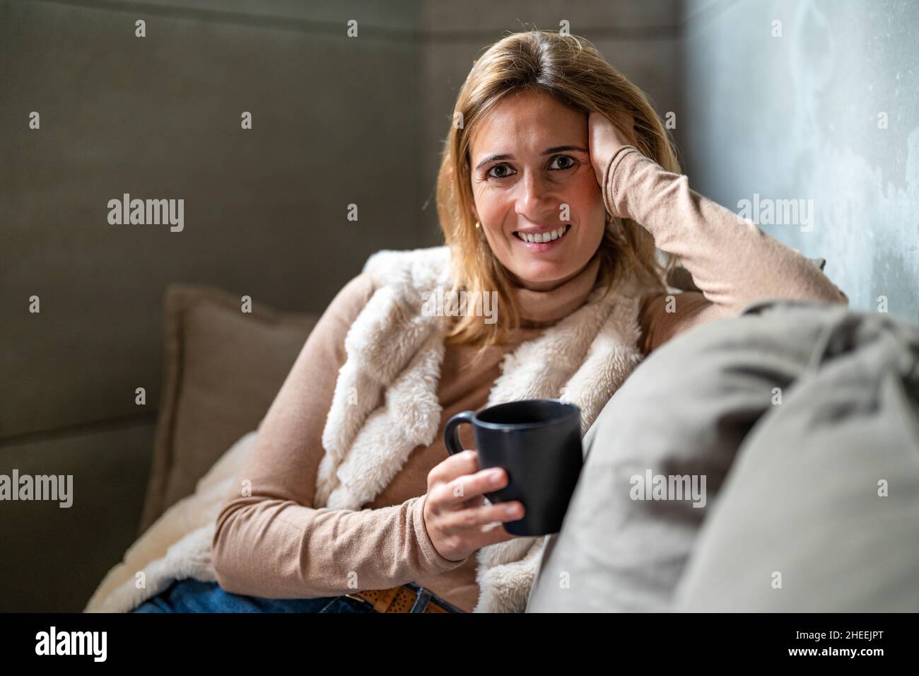 Lavoratore femminile positivo con una tazza di bevanda calda guardando la macchina fotografica mentre si siede sul divano con cuscini in ufficio durante la pausa Foto Stock