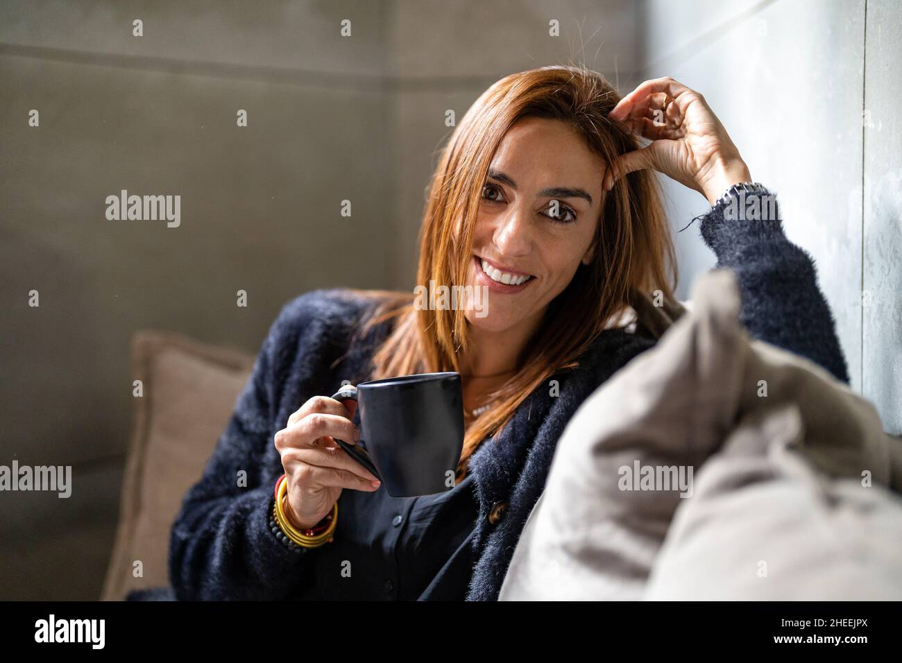 Lavoratore femminile positivo con una tazza di bevanda calda guardando la macchina fotografica mentre si siede sul divano con cuscini in ufficio durante la pausa Foto Stock